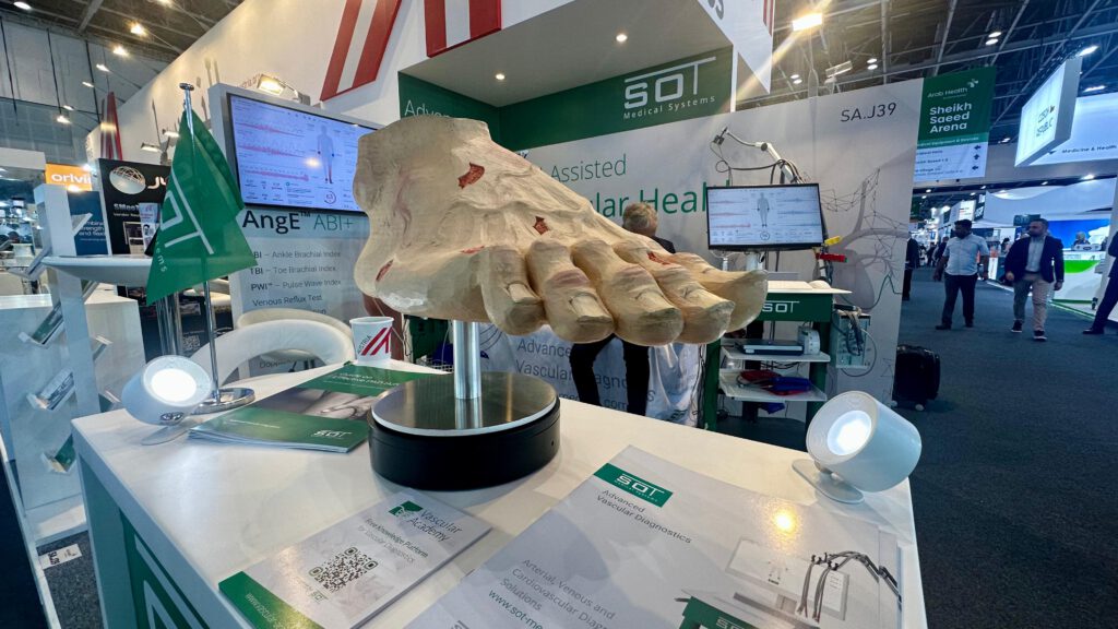 Ein Stand von SOT Medical Systems auf der Arab Health 2025, auf dem ein großes anatomisches Modell eines Fußes ausgestellt ist. Im Hintergrund sind Banner mit Informationen zum AngE™ ABI+ System sowie ein Bildschirm mit vaskulären Diagnosedaten zu sehen. Broschüren und eine grüne SOT-Flagge befinden sich auf dem Tisch.